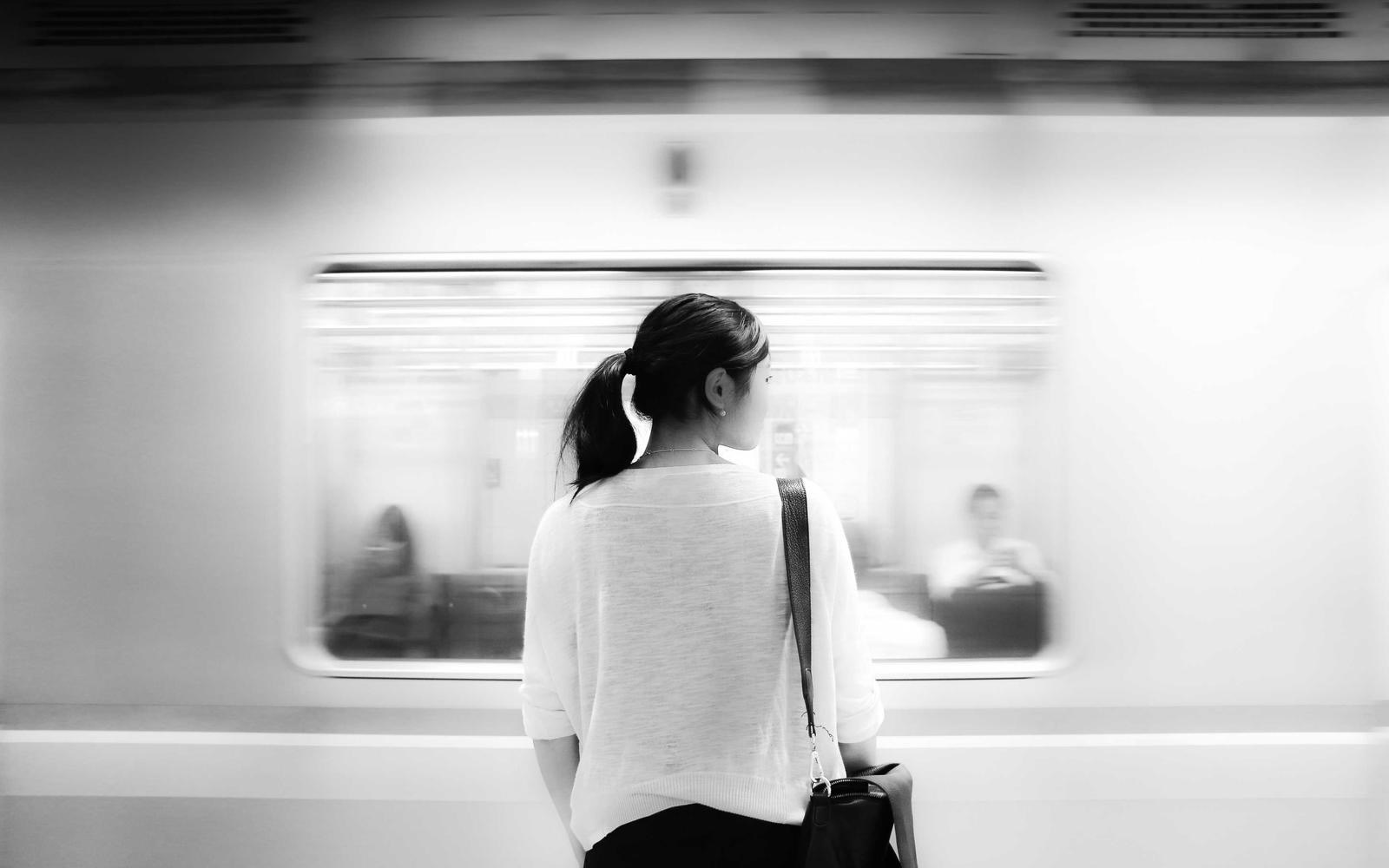 A woman waiting on a platform as a train passes, in black and white